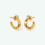 A pair of gold crescent-shaped hoop earrings with a small round clear gemstone at the top, displayed on a clean white background.