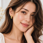 A close-up portrait of a woman with long brown hair wearing a small gold hoop earring with clear stones, resting her hand on her chin.