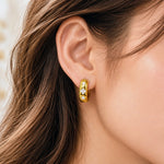 A close-up of a woman's ear wearing a small gold huggie hoop earring with three clear gemstones set in a row.