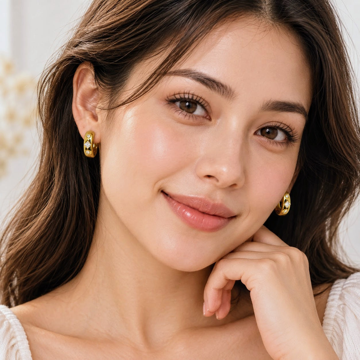 A close-up portrait of a woman wearing small gold hoop earrings with clear gemstone accents. The image is brightly lit, sharp, and has a soft, clean background.