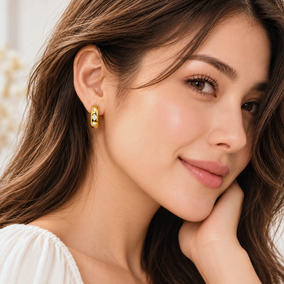A close-up of a woman wearing a small gold hoop earring with a textured, organic raised pattern. She has long brown hair and is wearing a white top, posing with her hand near her chin.