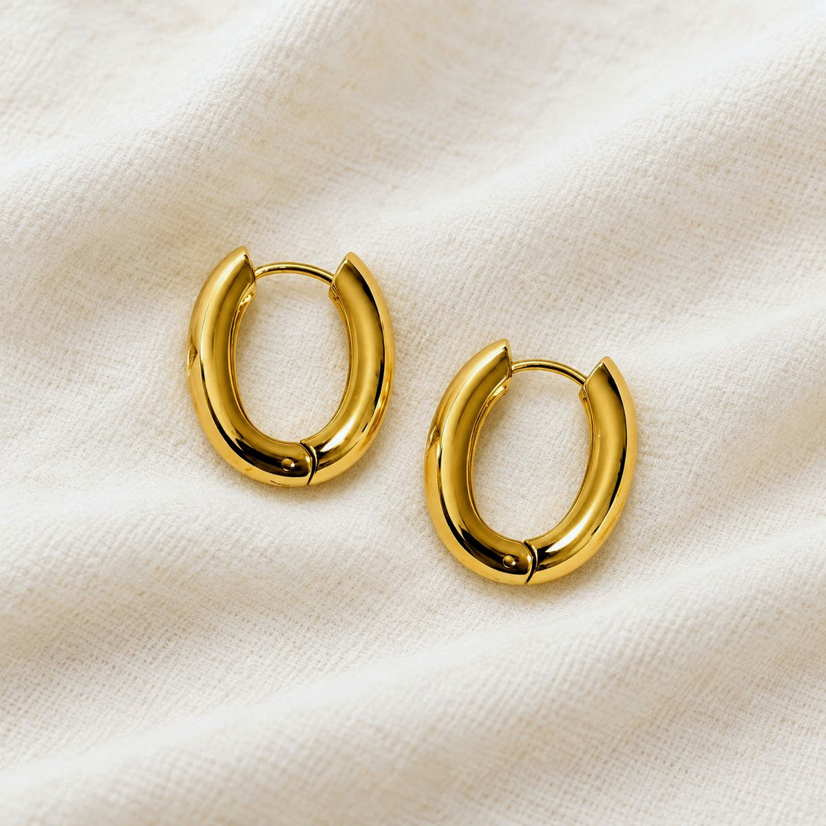 A pair of polished gold huggie hoop earrings resting on a textured, off-white fabric background.