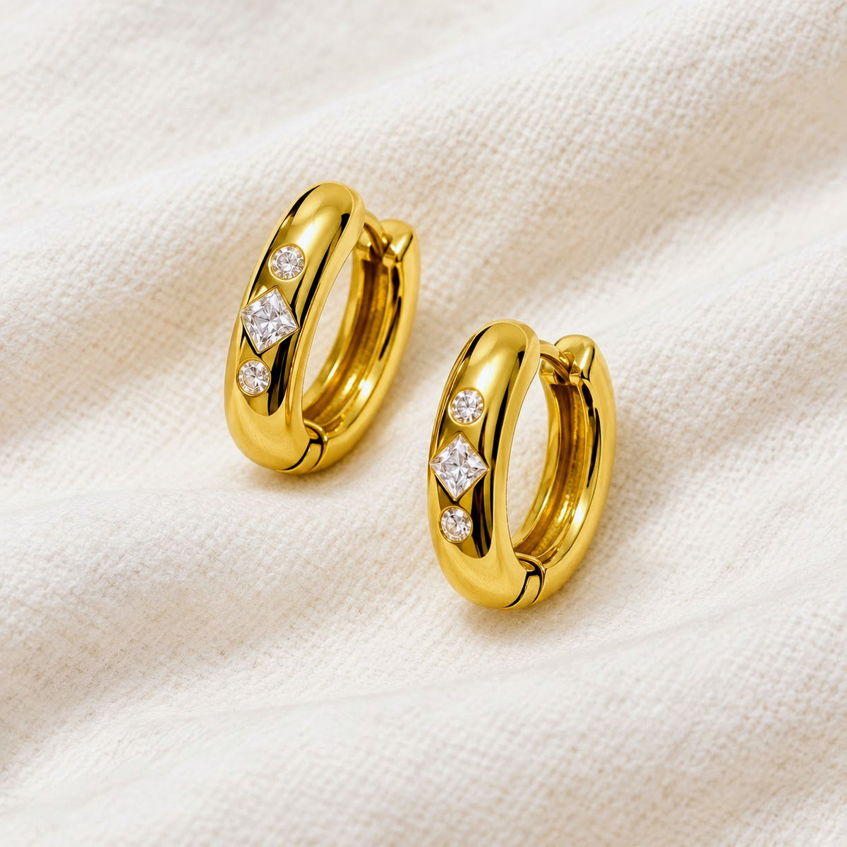 A pair of polished gold huggie hoop earrings, each adorned with three small clear gemstones, displayed on a textured off-white fabric background.