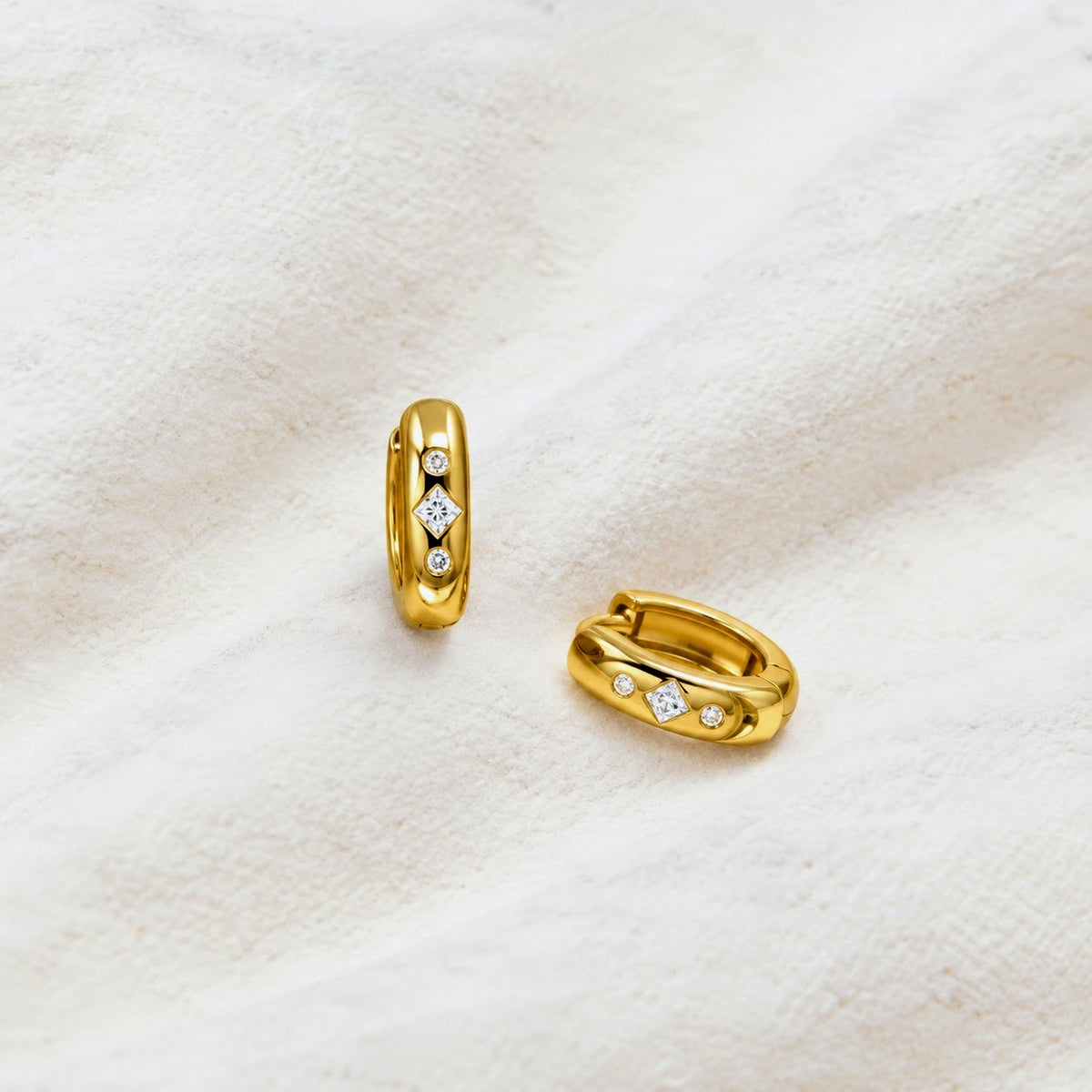 A pair of gold huggie hoop earrings with small clear gemstones set along the front, displayed on a soft, textured off-white fabric background.