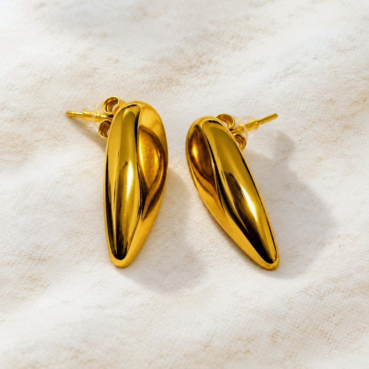 A pair of shiny gold teardrop-shaped stud earrings resting on a textured beige fabric background.