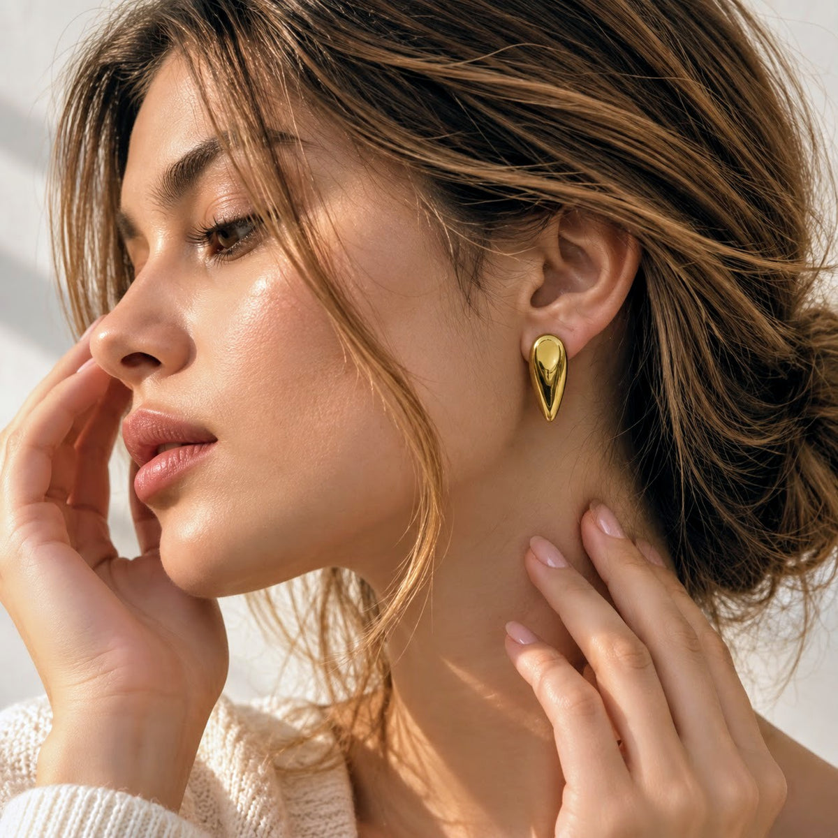 A close-up lifestyle shot of a woman wearing a gold teardrop-shaped stud earring, with her hands resting near her face against a bright, softly lit background.