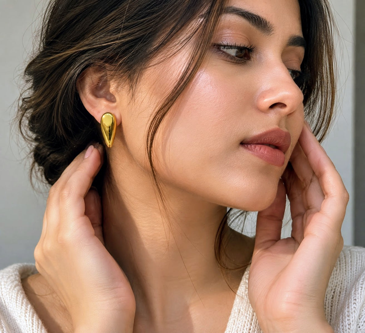 A close-up of a woman wearing a small, polished gold teardrop-shaped stud earring, with her hands gently touching her face and neck.