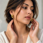 A close-up lifestyle shot of a woman wearing gold teardrop-shaped stud earrings, touching her earlobes. She has dark brown hair and is wearing a cream-colored knit top against a soft, light gray background.