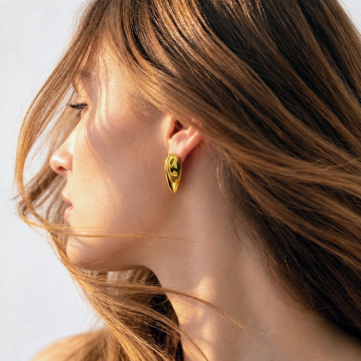 A close-up side profile of a woman with long light brown hair wearing a small, polished gold teardrop-shaped earring.