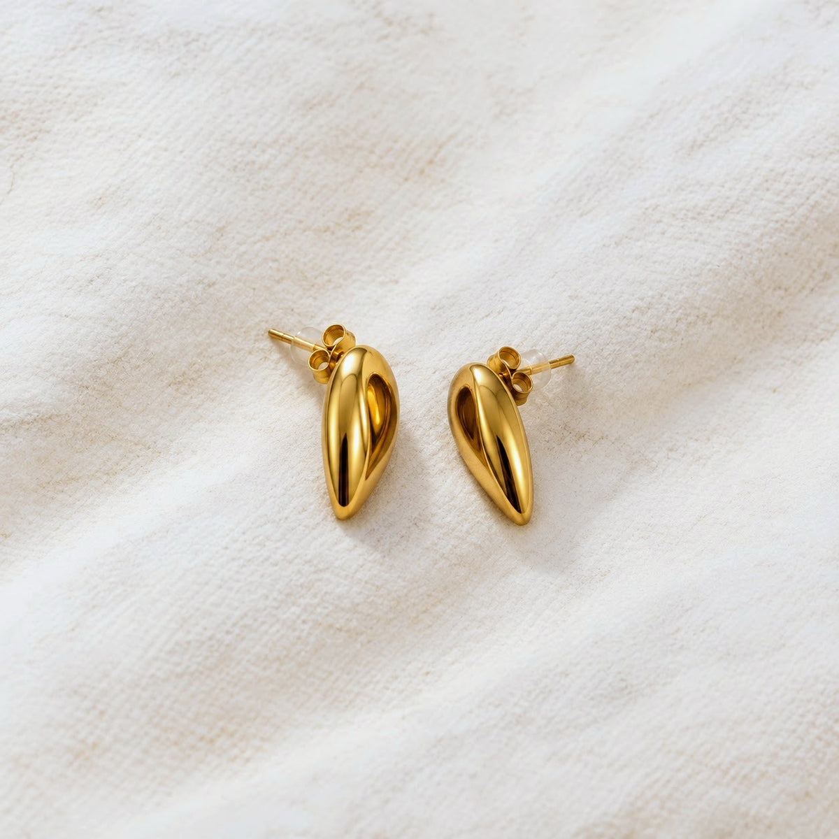 A pair of polished gold teardrop-shaped stud earrings resting on a textured, off-white fabric background.