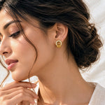 A close-up lifestyle shot of a woman wearing a gold hoop earring adorned with small clear stones and three dangling gold charms, set against a bright, softly lit background.