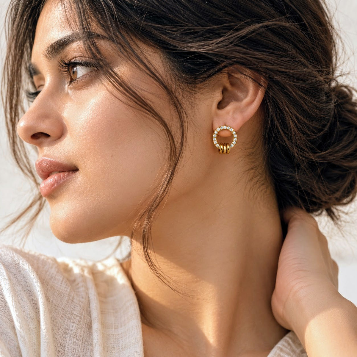 A close-up lifestyle shot of a woman wearing a gold hoop earring adorned with small white stones, shown in profile against a bright, softly lit background.