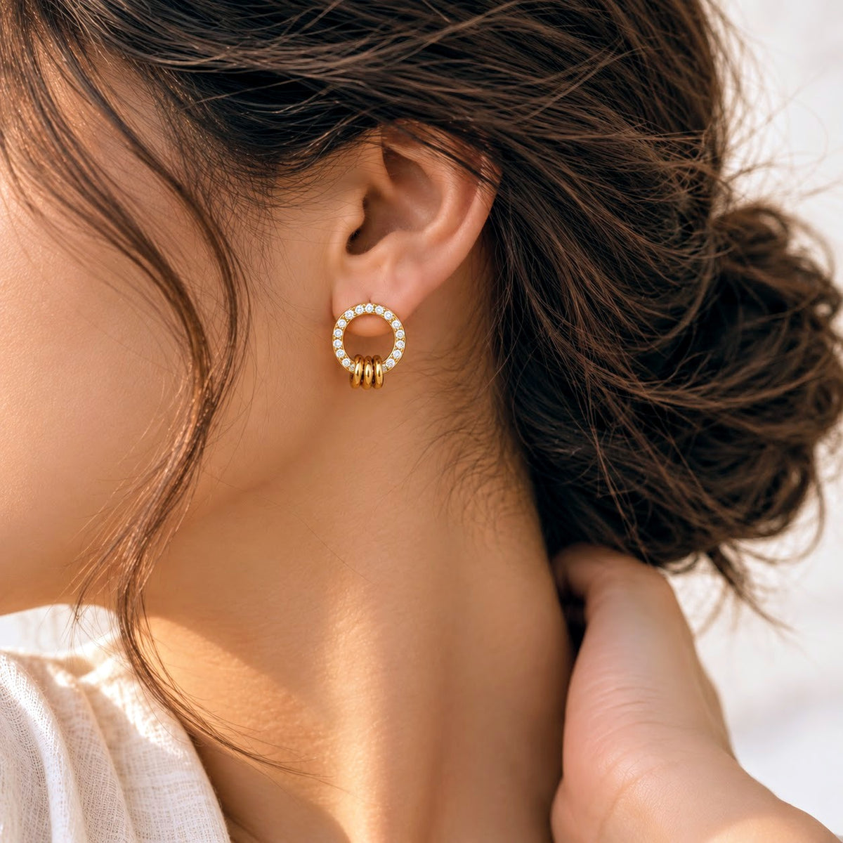 A close-up of a woman's ear wearing a gold hoop earring adorned with small clear stones and three dangling gold beads, against a soft light background.