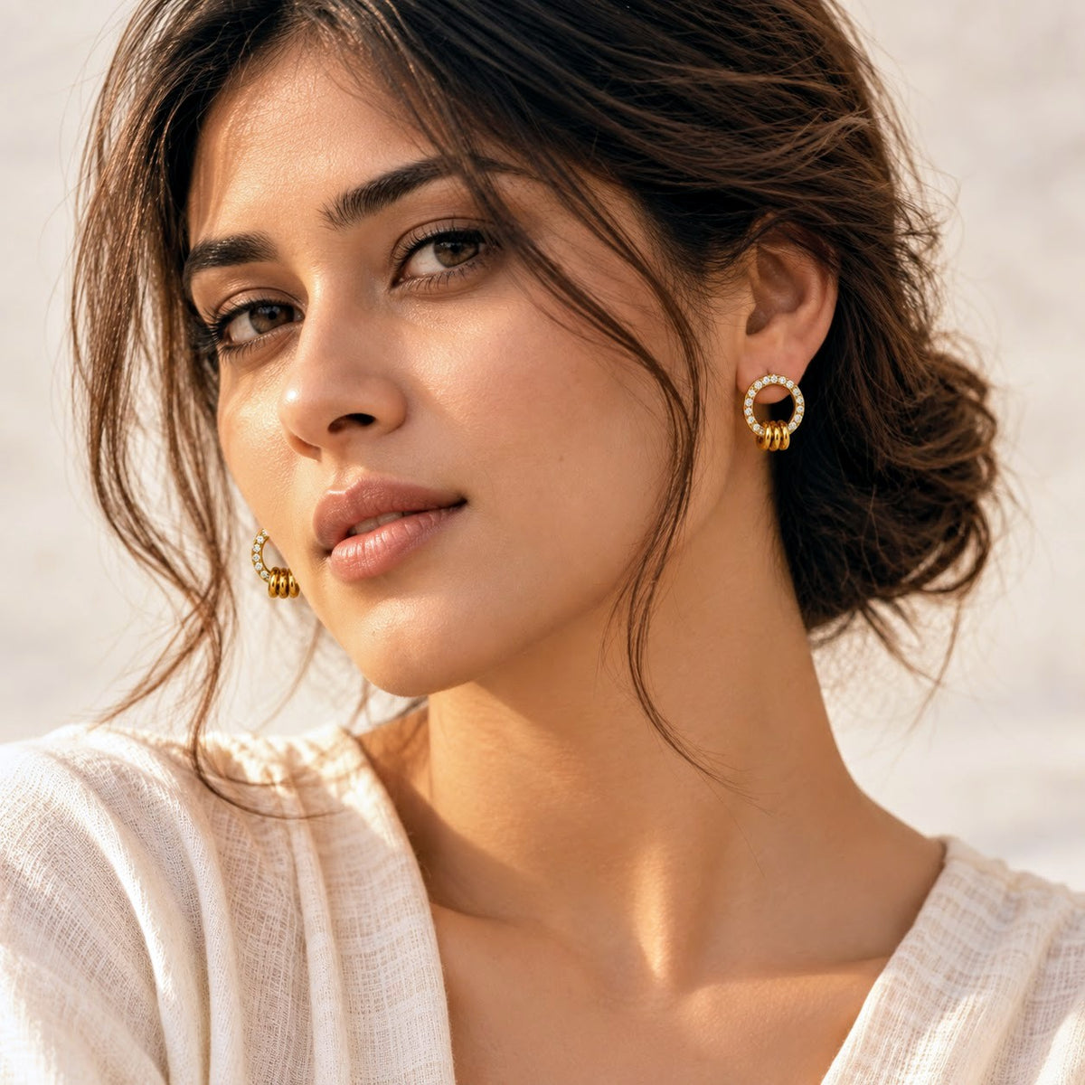 A close-up lifestyle shot of a woman wearing a gold hoop earring adorned with small white stones, shown in profile against a bright, softly lit background.