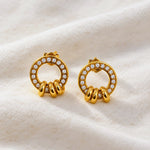A pair of gold hoop earrings with clear crystal accents and three twisted gold barbell details at the bottom, displayed on a soft, textured cream fabric background.