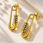 A pair of gold safety-pin stud earrings with polished silver bead accents, displayed on a textured beige surface with soft shadows.