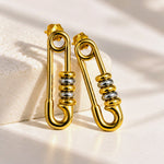 A pair of gold safety-pin style hoop earrings with silver bead accents, displayed on a textured white block with soft shadows.