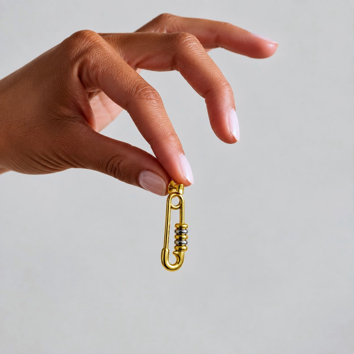 A close-up of a hand holding a small gold safety-pin-style charm or accessory against a plain warm beige background.