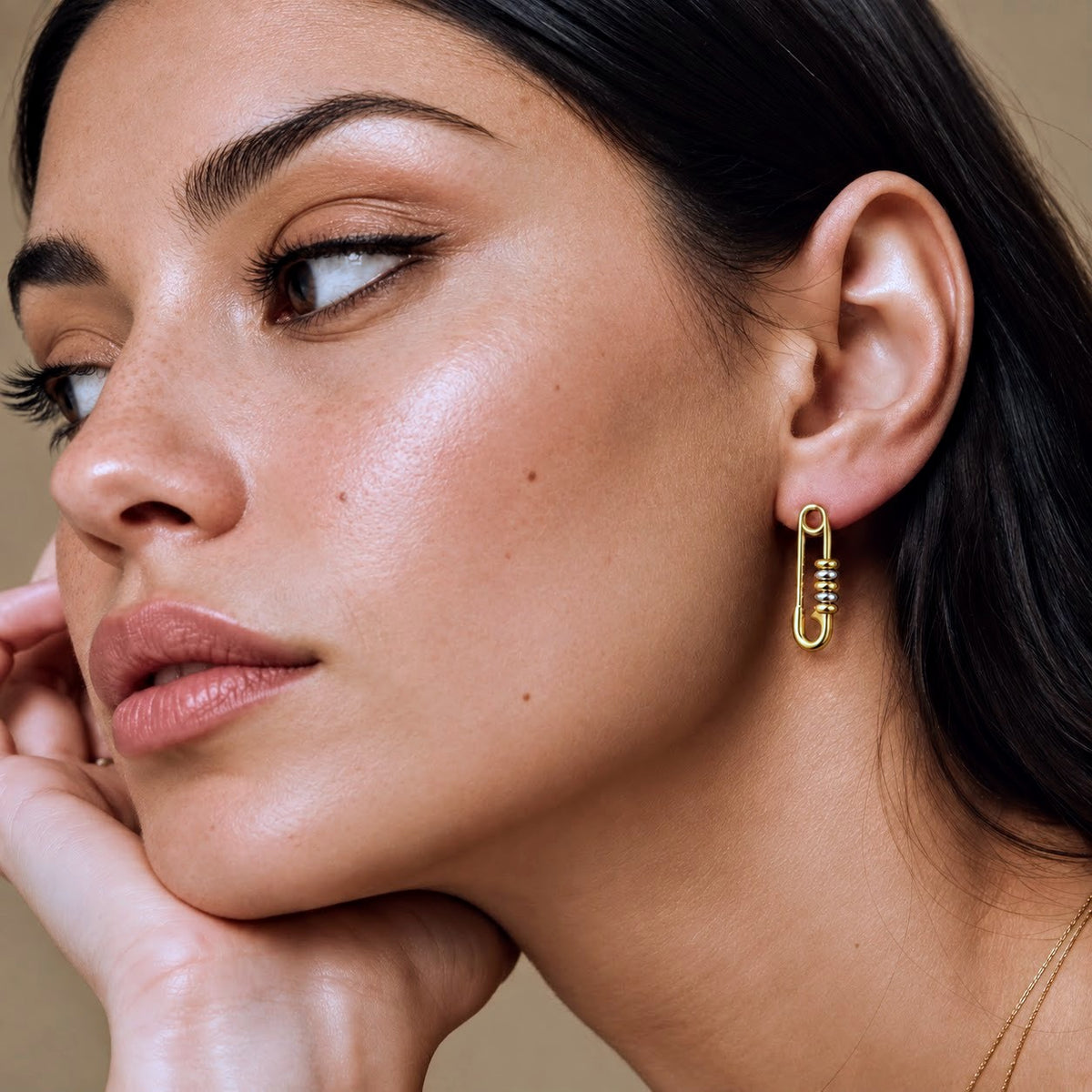 A close-up of a woman wearing a delicate gold safety-pin earring with small clear stones, set against a soft beige background.