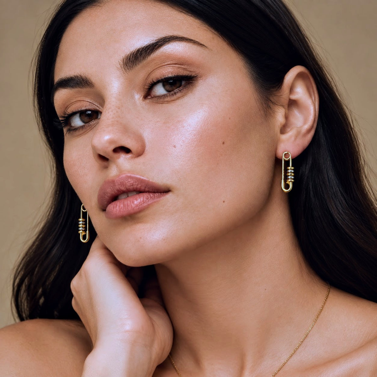 Close-up of a woman wearing gold safety-pin drop earrings and a delicate gold necklace against a warm beige background.
