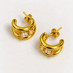 A pair of gold crescent-shaped hoop earrings with clear gemstones, displayed on a clean off-white background.