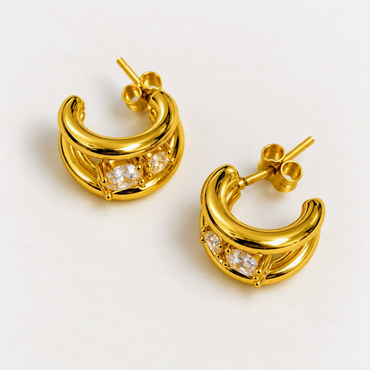 A pair of gold crescent-shaped hoop earrings with clear gemstones, displayed on a clean off-white background.