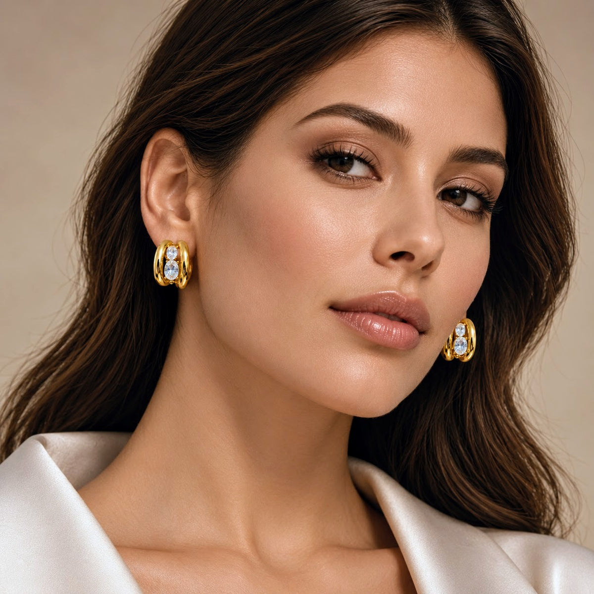 A close-up portrait of a woman wearing gold hoop earrings with clear gemstones, set against a soft beige background and a white satin collar.
