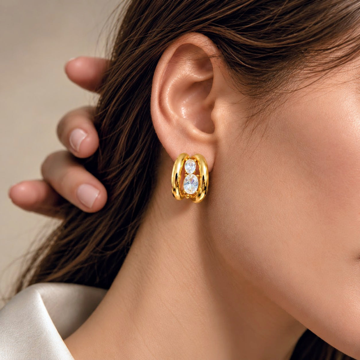 A close-up of a model's ear wearing a gold huggie hoop earring with three round clear gemstones, set against a soft beige background.