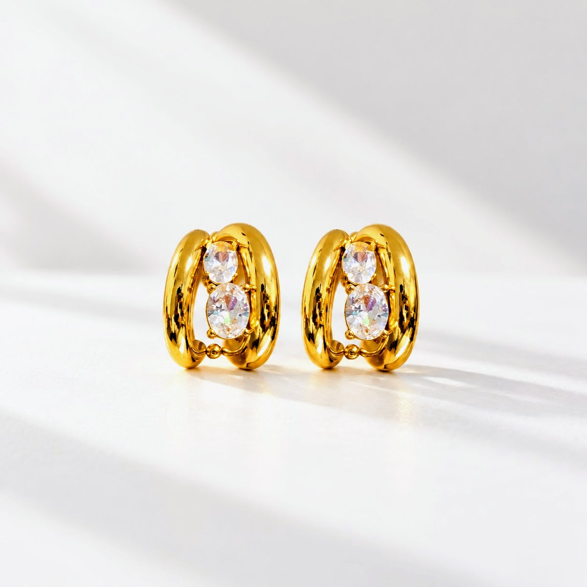 A pair of gold huggie hoop earrings with three clear oval gemstones on each earring, displayed on a clean white surface with soft diagonal shadows.
