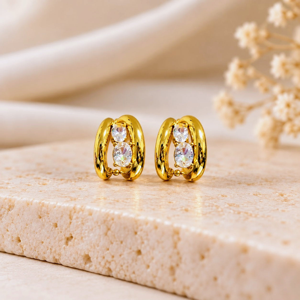 A pair of gold hoop-style stud earrings with clear oval gemstones, displayed on a light beige stone slab with soft cream fabric and dried flowers in the background.