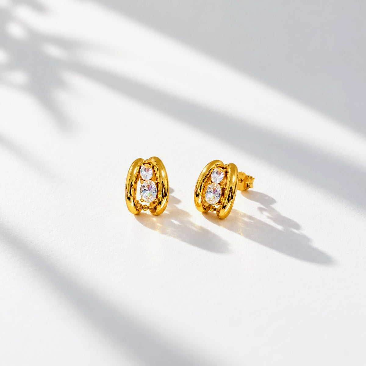 A pair of gold-toned twisted hoop stud earrings with clear oval gemstones, displayed on a clean white surface with soft leaf shadows.