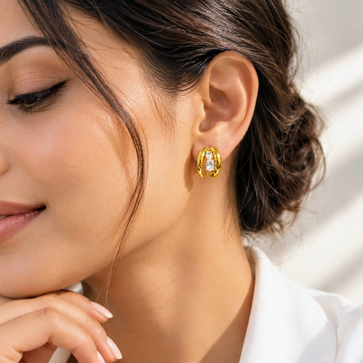 A close-up lifestyle shot of a woman wearing a gold hoop earring with a central clear gemstone, set against a bright, softly lit background.
