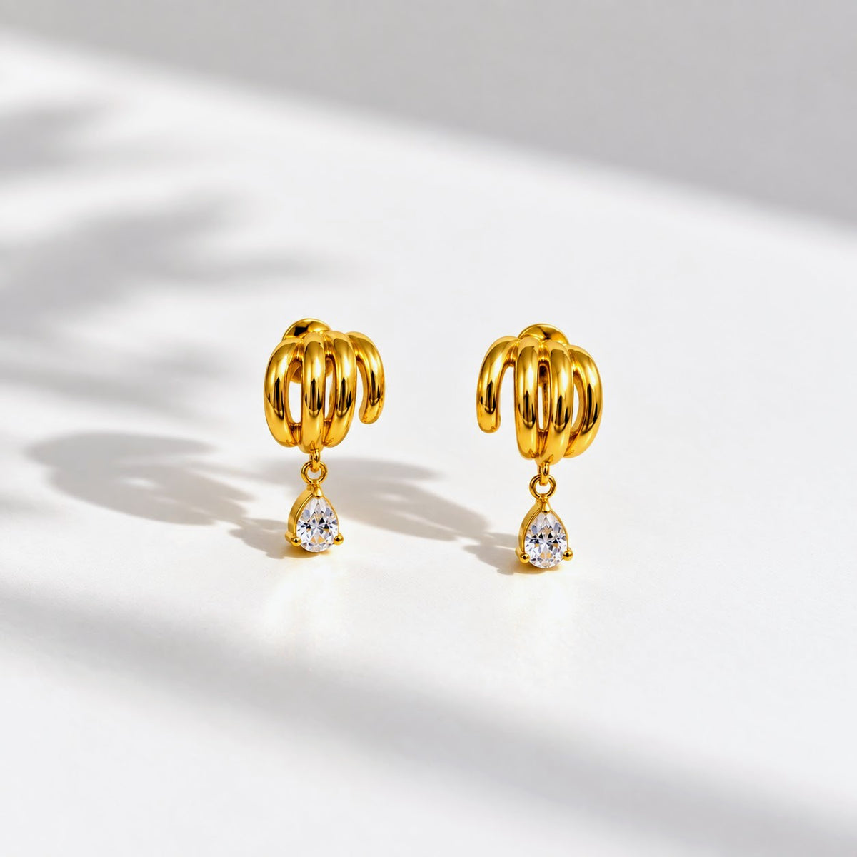 A pair of gold-toned dangle earrings with a textured, ribbed top and a small clear teardrop gemstone hanging below, displayed on a white surface with soft shadows.