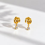 A pair of gold-toned dangle earrings with a textured, ribbed top and a small clear teardrop gemstone hanging below, displayed on a white surface with soft shadows.