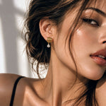 A close-up lifestyle shot of a woman wearing a gold dangle earring with a clear teardrop gemstone, set against a bright, sunlit background.