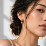 A close-up of a woman wearing a gold dangle earring with a clear teardrop gemstone, shown in profile against a softly lit white background.