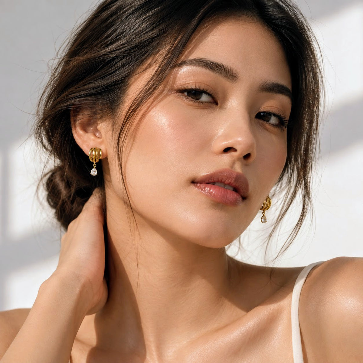 A close-up lifestyle shot of a woman wearing delicate gold drop earrings with small clear teardrop crystals, set against a bright, softly shadowed white background.