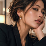 A close-up portrait of a woman wearing a delicate gold dangle earring with a small clear teardrop stone, resting her head on her hand.
