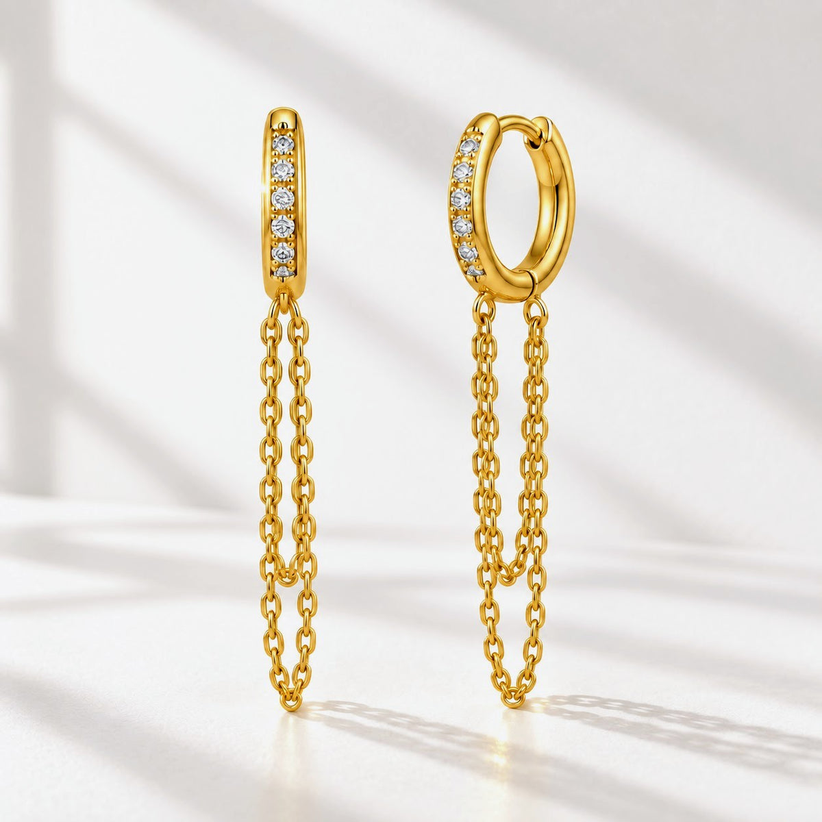 A pair of gold huggie hoop earrings with a vertical bar of small clear stones on one side and a dangling double-chain design on the other, set against a bright white background with soft diagonal shadows.