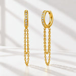 A pair of gold huggie hoop earrings with a vertical bar of small clear stones on one side and a dangling double-chain design on the other, set against a bright white background with soft diagonal shadows.