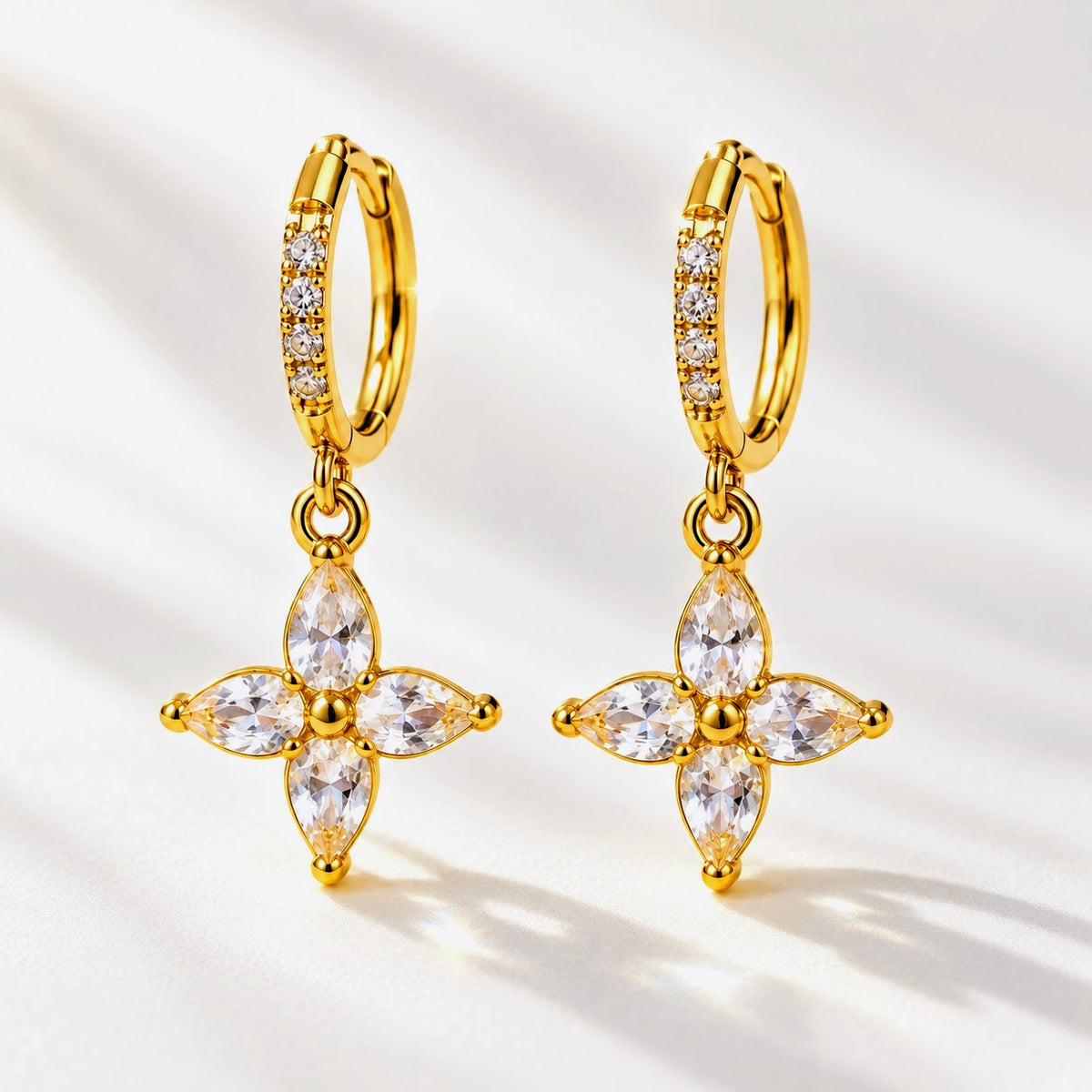 A pair of gold huggie hoop earrings with dangling floral cross charms, set with clear marquise and round-cut stones, displayed on a clean white background with soft shadows.