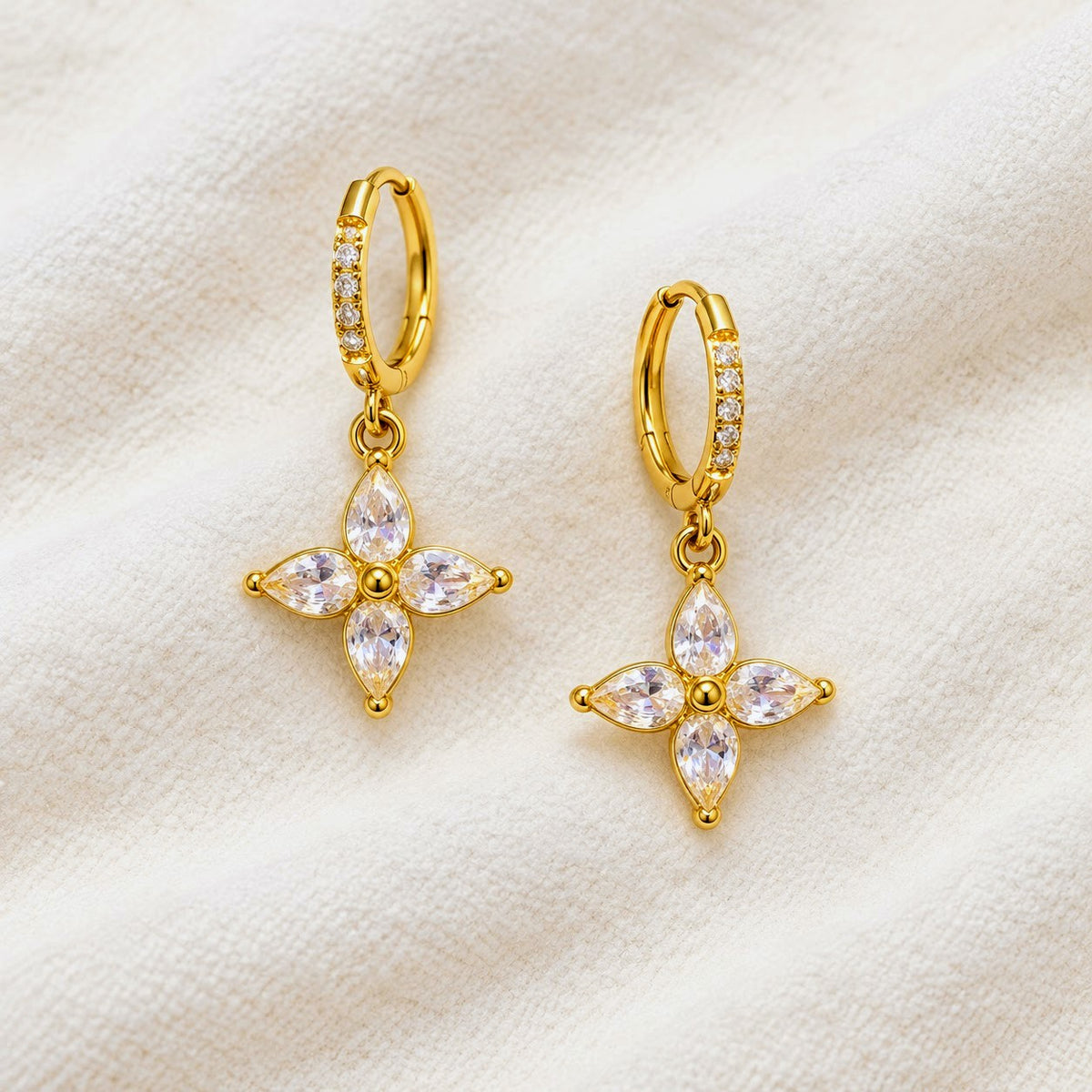 A pair of gold huggie hoop earrings with dangling four-petal floral charms set with clear marquise and round gemstones, displayed on a soft white textured fabric background.