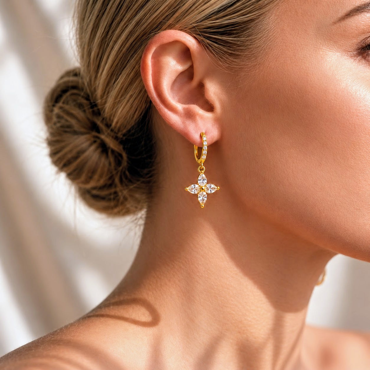 A close-up of a woman's ear and neck wearing a gold huggie hoop earring with a dangling four-petal flower charm set with clear stones.