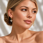 A close-up of a woman wearing gold dangle earrings with clear gemstones, shown against a softly lit neutral background with leaf shadows.