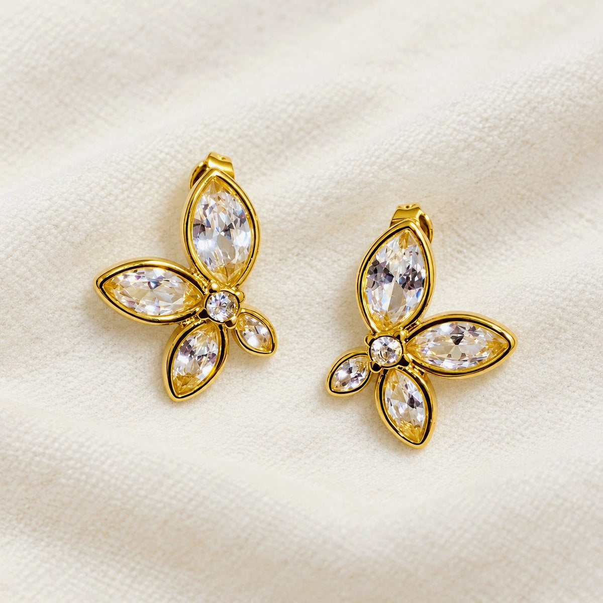 A pair of gold butterfly-shaped earrings with clear marquise and round crystals, displayed on a textured cream fabric background.