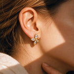 A close-up of a woman's ear wearing a small gold flower-shaped stud earring with clear gemstones, set against warm sunlight and shadow.