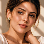 A close-up portrait of a woman wearing small gold butterfly-shaped stud earrings with clear stones, set against a warm beige background with soft shadows.