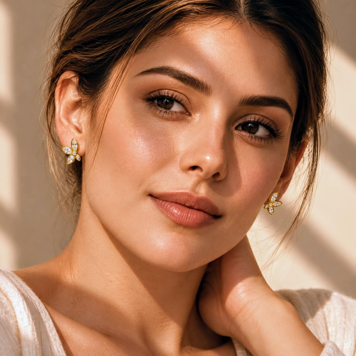A close-up portrait of a woman wearing small gold butterfly-shaped stud earrings with clear stones, set against a warm beige background with soft shadows.
