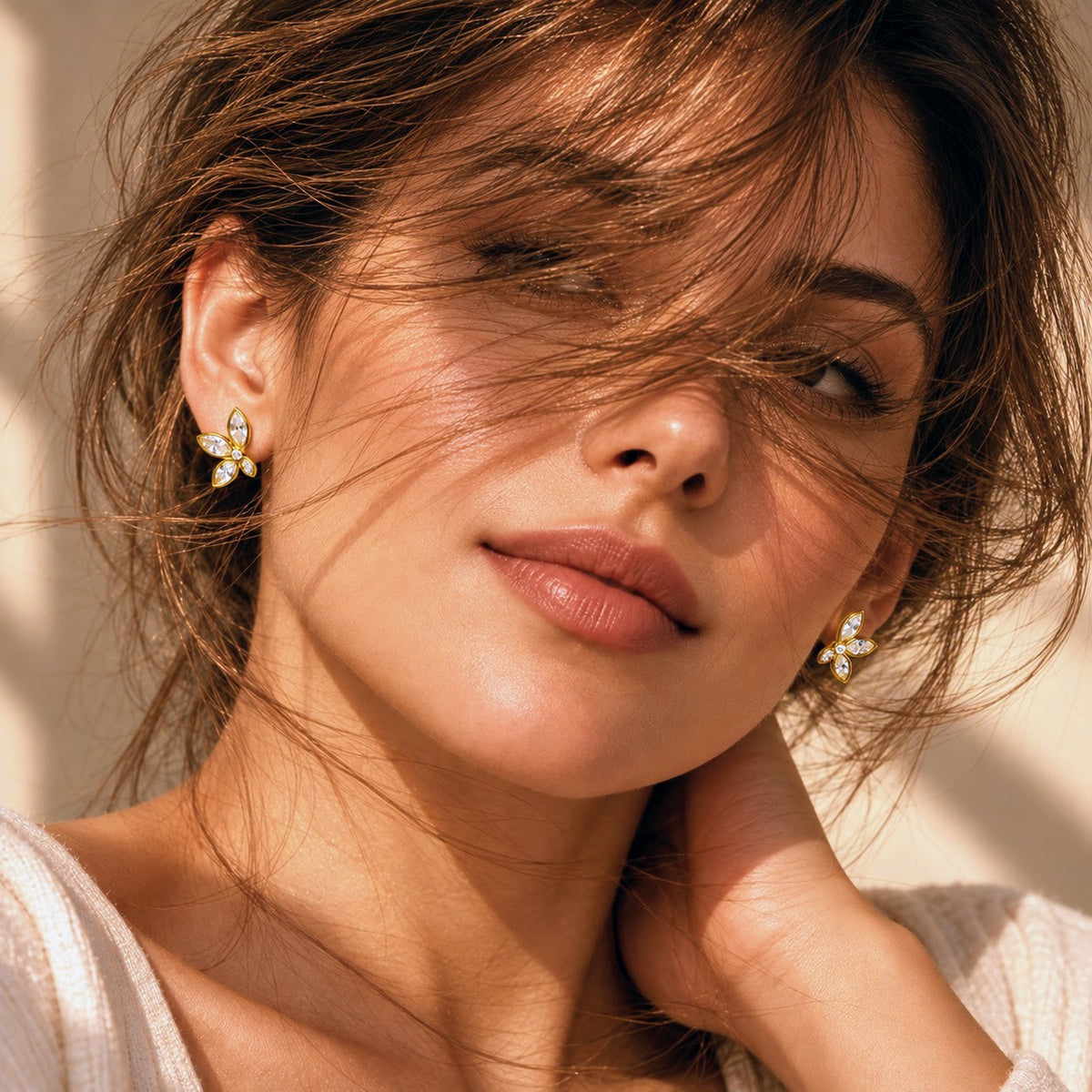 A close-up lifestyle shot of a woman with windblown hair wearing small gold flower-shaped stud earrings with clear stones.