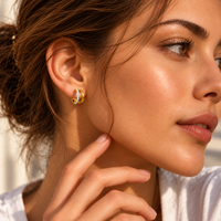A close-up lifestyle shot of a woman wearing a small gold hoop earring with a row of clear stones, shown in warm natural sunlight.